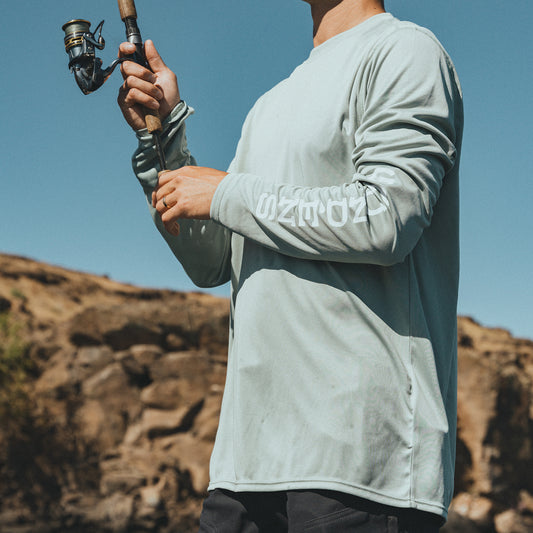 Person wearing light gray Grundens long sleeve shirt while holding a fishing rod with a scenic background
