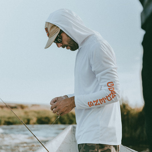 Man wearing a white hoodie with red Grundens text, fishing by a body of water.