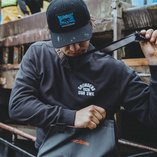 Person wearing a dark hoodie and cap with 'Grundéns' branding, adjusting a belt.