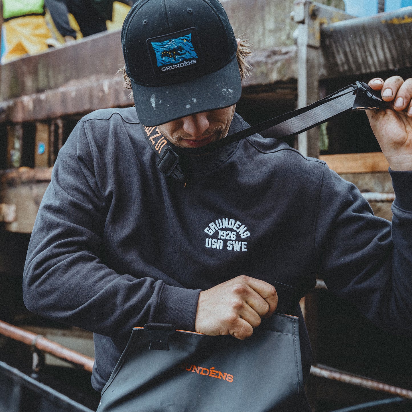 Person wearing a dark hoodie and cap with 'Grundéns' branding, adjusting a belt.