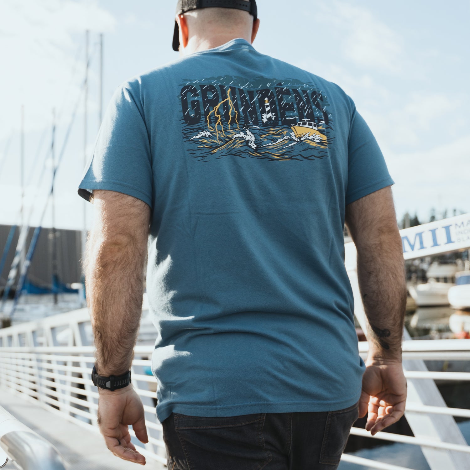 Man wearing a blue t-shirt with a Grundens graphic design on the back, standing on a boat.