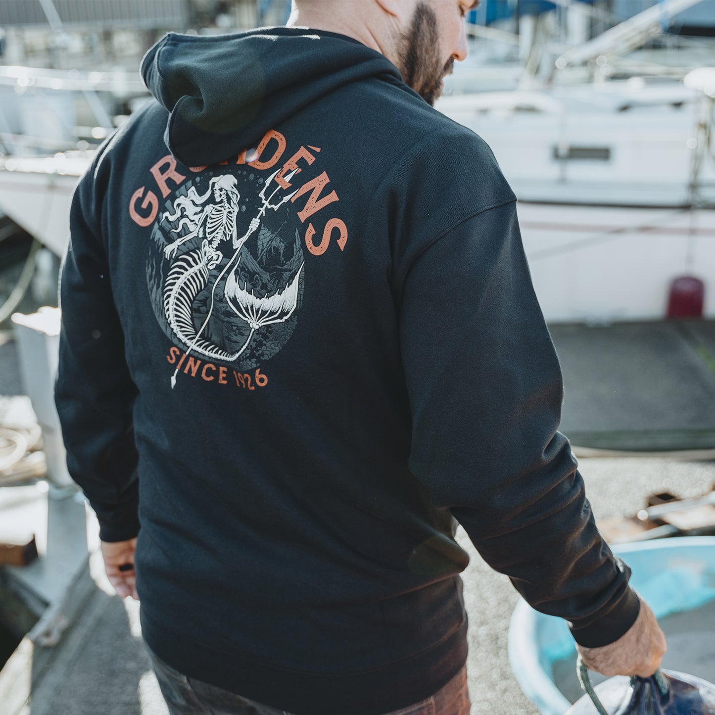 Man wearing a black hoodie with Grundens brand logo on the back, standing near boats.