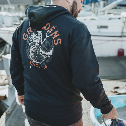 Man wearing a black hoodie with Grundens brand logo on the back, standing near boats.