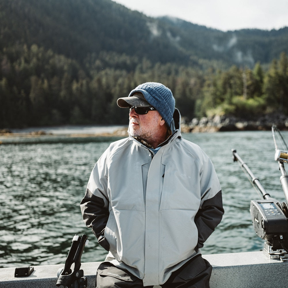 Man wearing a light gray GORE-TEX® jacket and blue hat, standing on a boat near a forested shoreline.